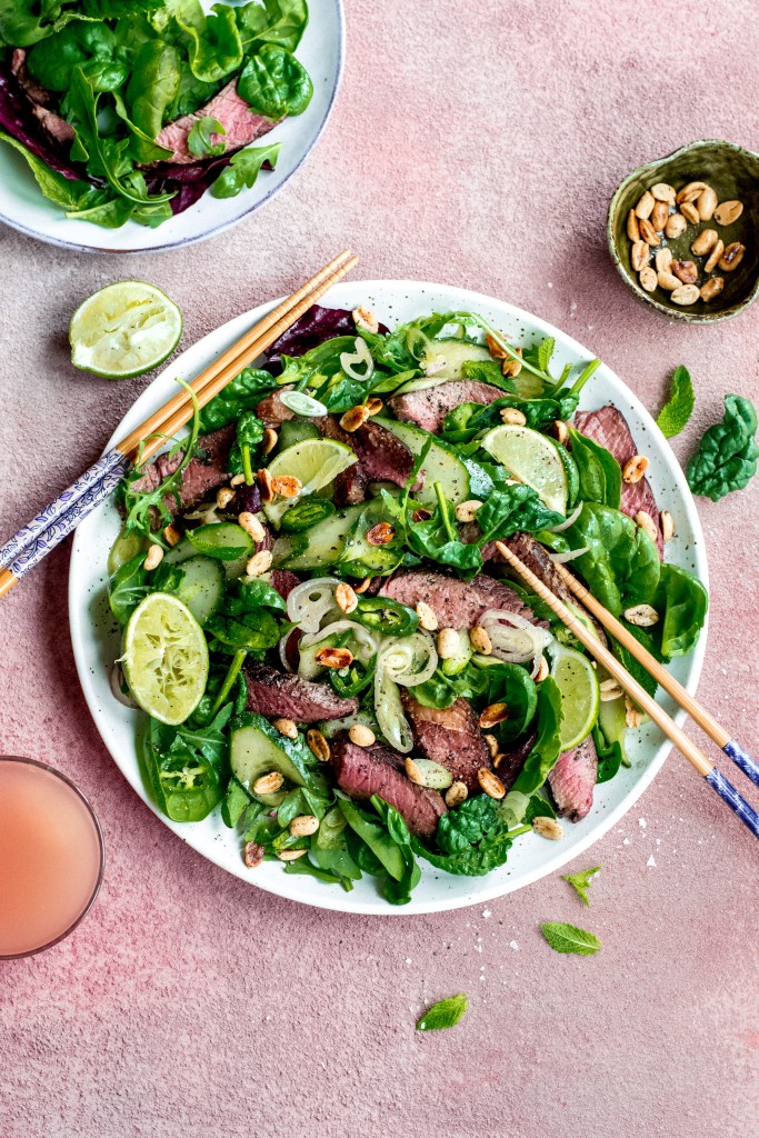 Thai-inspired beef salad. Fresh mixed greens, Irish beef, peanuts, shallots, mint and lime on two plates - pink background. Recipe development, food photography and styling by Chiara Gianelli.