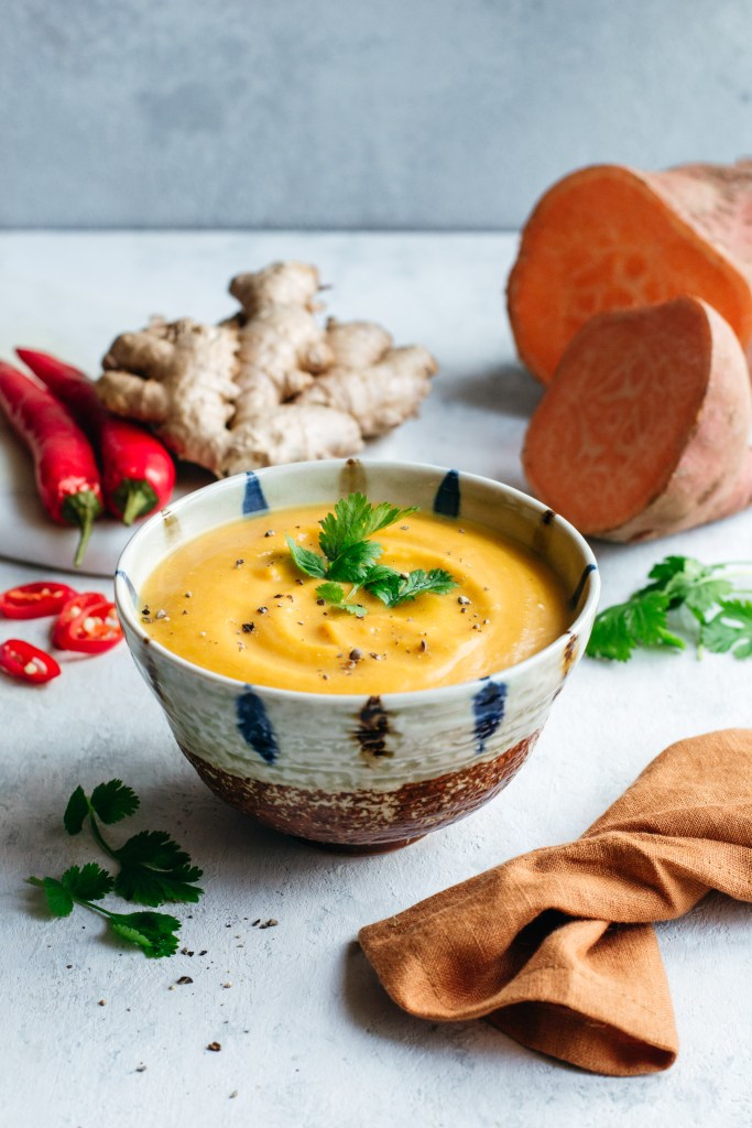 Sweet potato soup in a small bowl with the key ingredients (ginger, red chillies, coriander and sweet potato) in the background.
