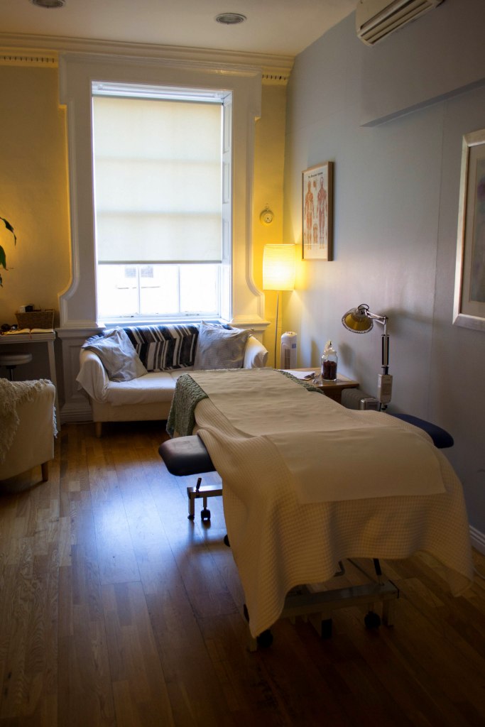 A calming therapy room featuring a massage table, a comfortable chair, and natural light filtering through a window, creating a serene atmosphere.