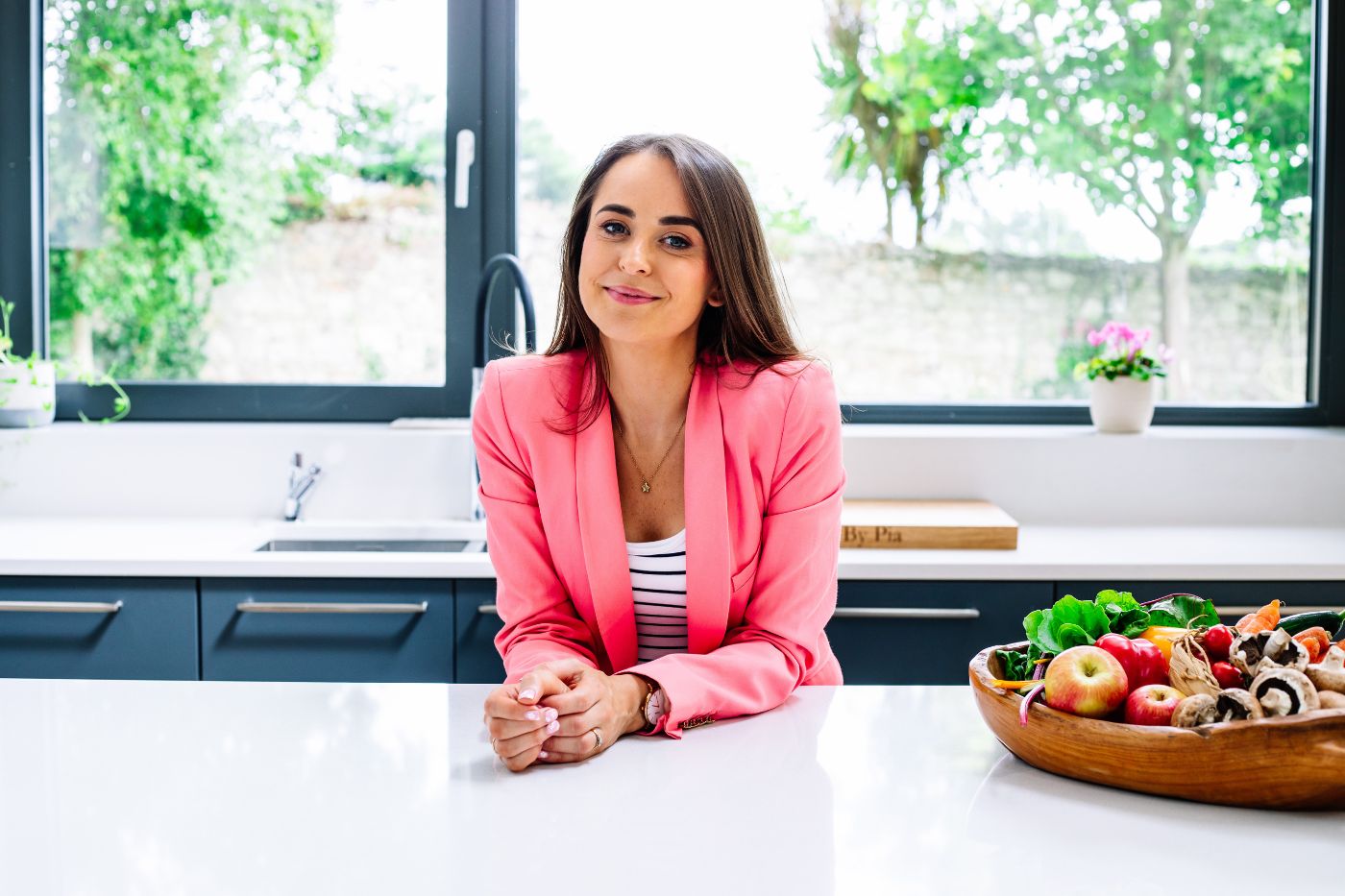 Woman wearing a pink blazer standing in front of a window in a kitchen. Dublin brand photography by Chiara Gianelli