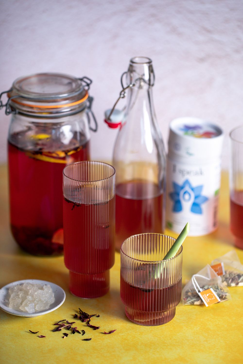 Glasses and bottles filled with red water kefir. Kefir grains in a small bowl, tea bags, and a package of tea. All on a yellow background.