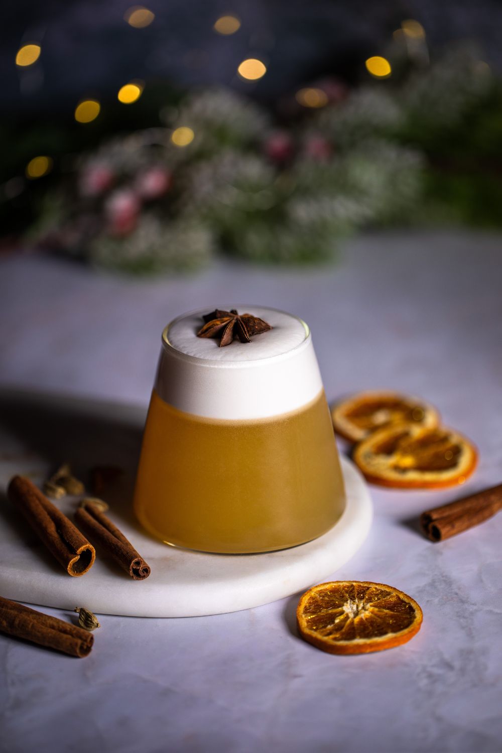 Glass with a drink made with egg whites and served with star anise on top. Around the glass there are dried orange slices, cinnamon, cardamon, and Christmas decorations. Styled by food photographer Chiara Gianelli in Dublin