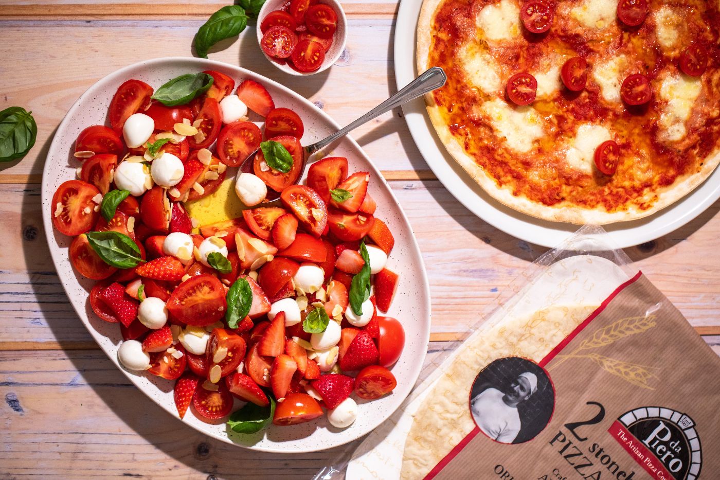 Top down photo of a salad with tomatoes, strawberries and mozzarella pearls. On the wooden table there is the salad, a margherita pizza, fresh tomatoes and a pizza base package. Dublin food photography by Chiara Gianelli