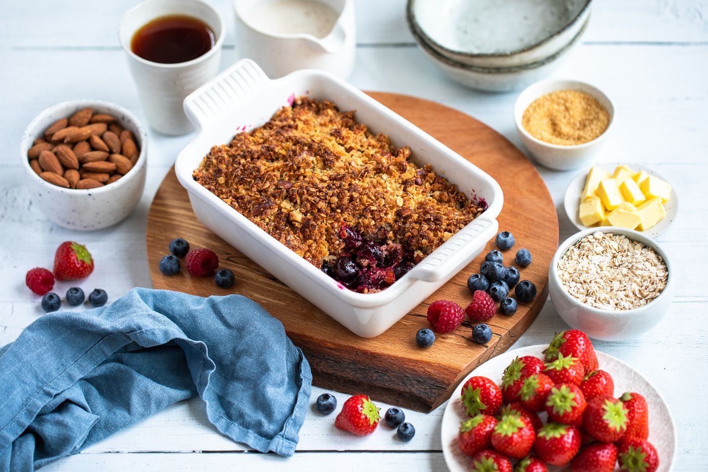 Crumble with berries placed on a wooden board. Around the board there are fresh berries, butter, oats, sugar, almonds, a cup of tea, a blue napkin and a pitcher with milk.