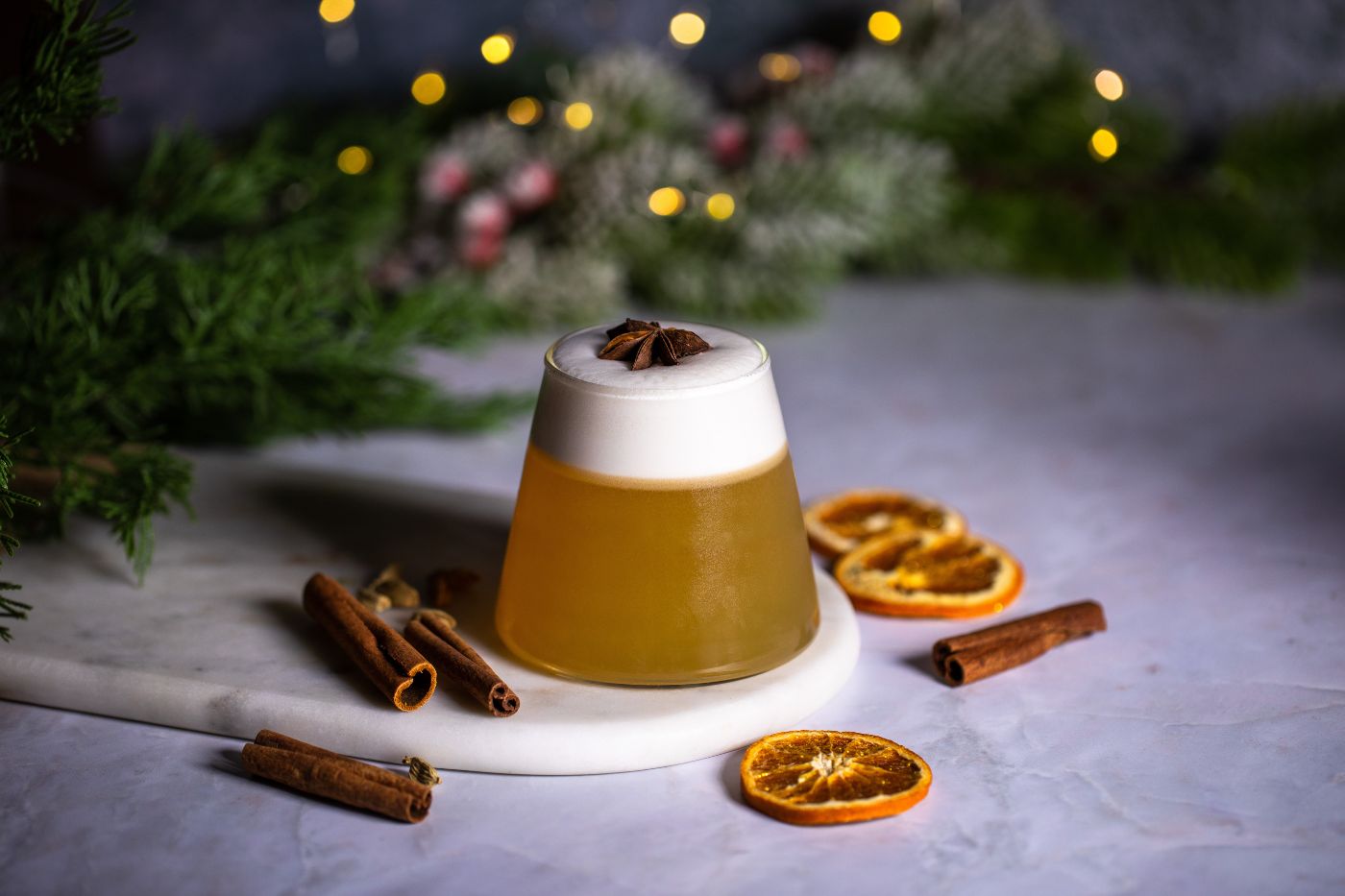 Glass with a whiskey sour and served with star anise on top. Around the glass there are dried orange slices, cinnamon, cardamon, and Christmas decorations. Styled by food photographer Chiara Gianelli in Dublin