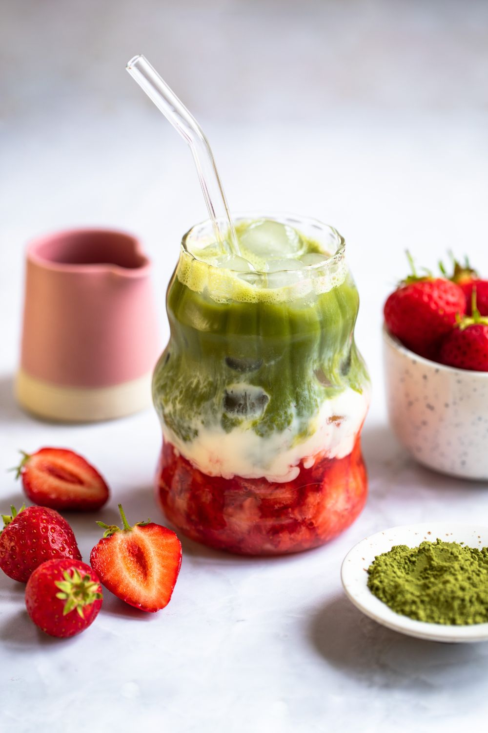 Glass of iced strawberry matcha with visible red, white and green layers. Fresh strawberries, a pink pitcher, and matcha powder around the glass. Food photography and styling by Chiara Gianelli in Ireland