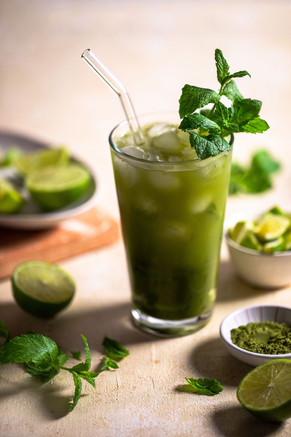 Glass with a matcha mojito served with ice and fresh mint. Around the glass there is mint, limes cut into pices, and matcha powder. Drink photo by photographer Chiara Gianelli