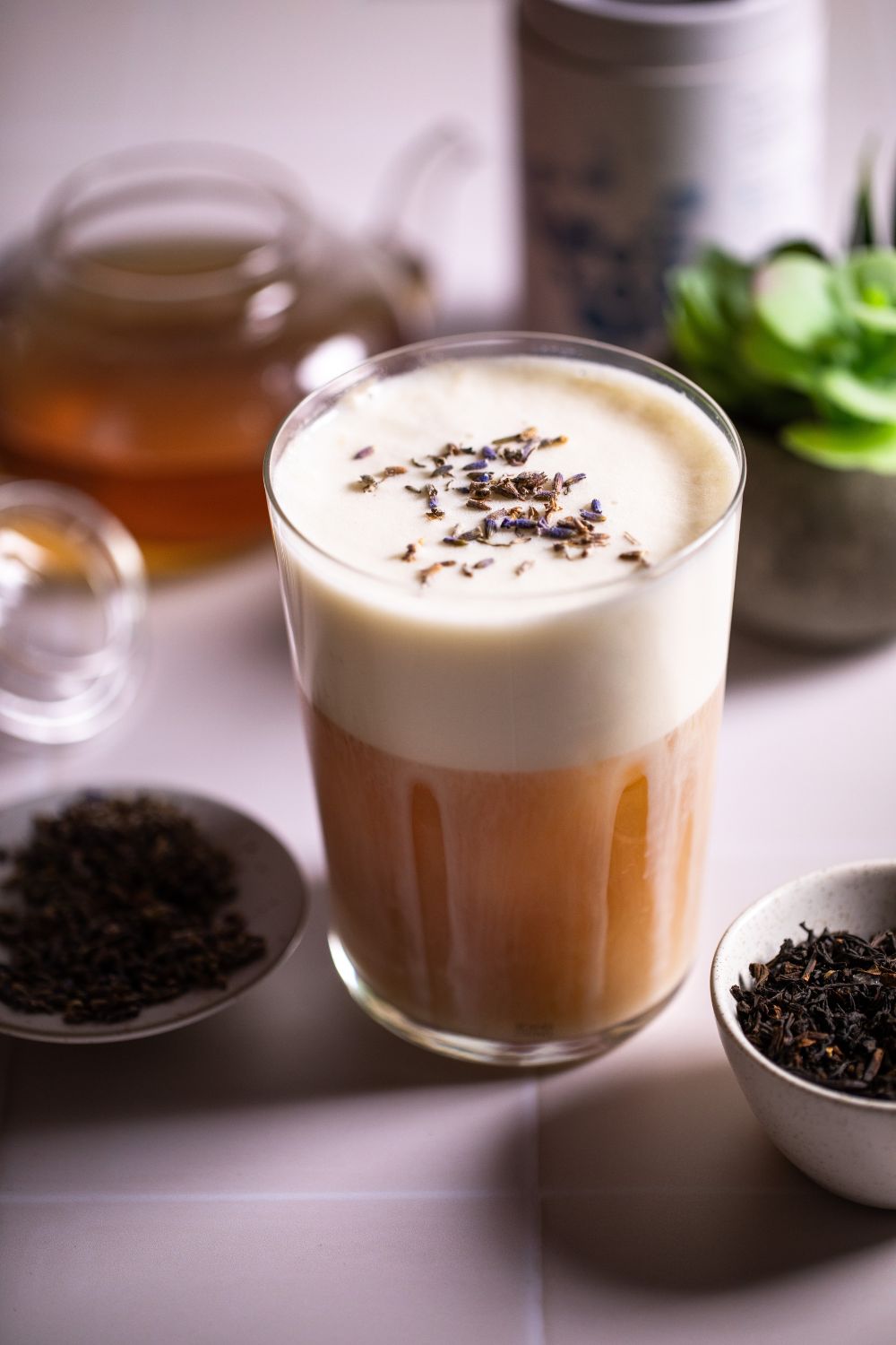 Glass with a non alcoholic London fog drink and dried lavander on top. Around the glass there is a teapot, a small plant, tea, lavander, and a tea package. Drink photography by Chiara Gianelli