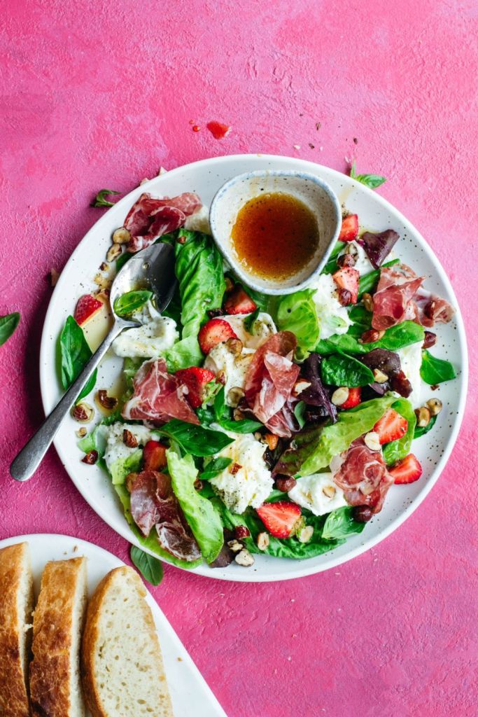 Salad with mixed greens, mozzarella, basil, strawberries, hazelnuts and coppa served with a sauce and bread, with the plate placed on a pink background. Styled by food photographer Chiara Gianelli in Ireland