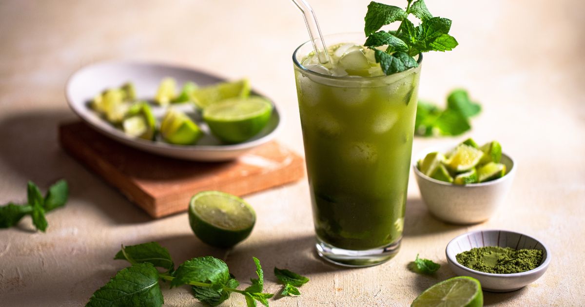 Matcha drink in a tall glass with fresh mint as a garnish. Around the glass, there are a small bowl with matcha powder, limes, and fresh mint. Food photo and styling by food photographer Chiara Gianelli