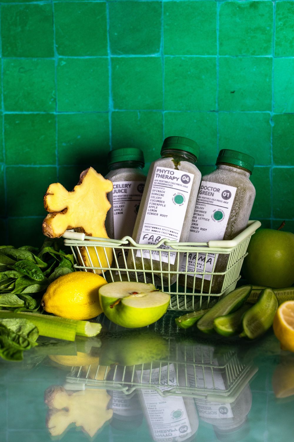 Three bottles of frozen green cold pressed juice placed in front of a background made of green tiles. The bottles are inside a small green shopping basket and around it there are fresh fruit and vegetables. Product photography by Chiara Gianelli